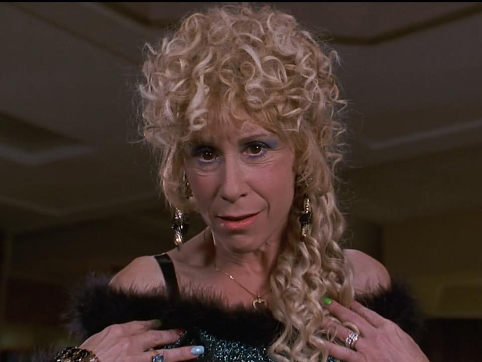 Rhea Perlman as Zinnia wearing a green dress with a fluffy black collar and with blonde, curly hair draping down her left shoulder