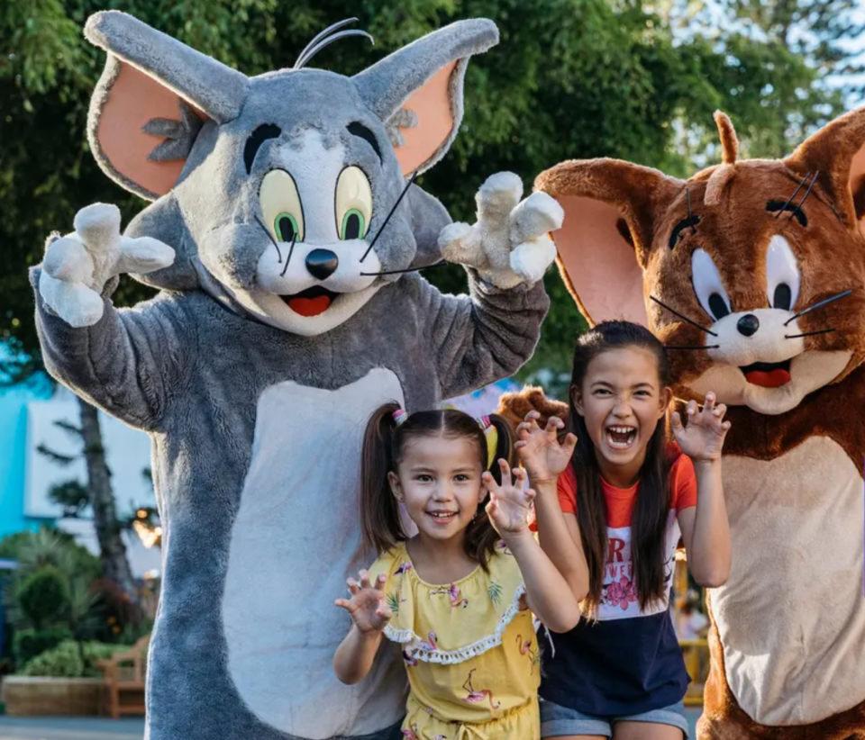 Oversize fluffy characters stand behind two girls with long dark hair wearing summery holiday clothing with greenery behind