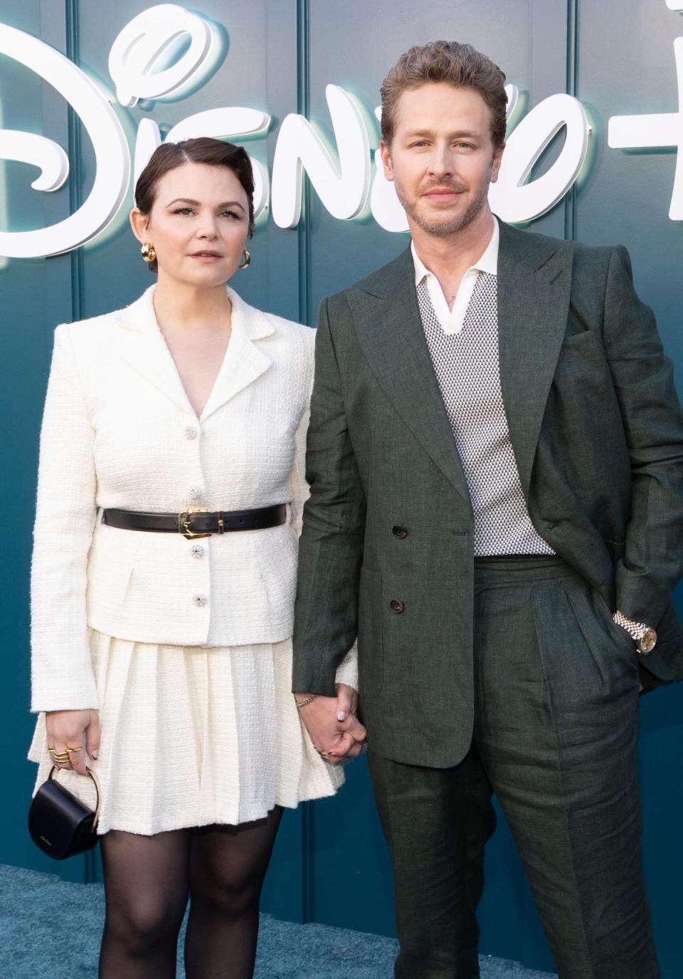 Two celebrities pose at a Disney+ event; one wears a white jacket and skirt combo, the other a green suit