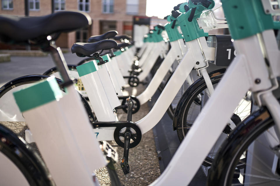 Modern electric rental bikes lined up during charging on a sunny day, identified by number, brick building in background.