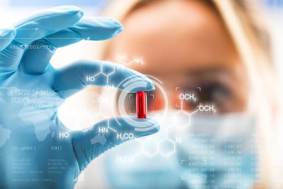 A woman wearing a medical face mask and gloves holds a medicine capsule in her fingers.