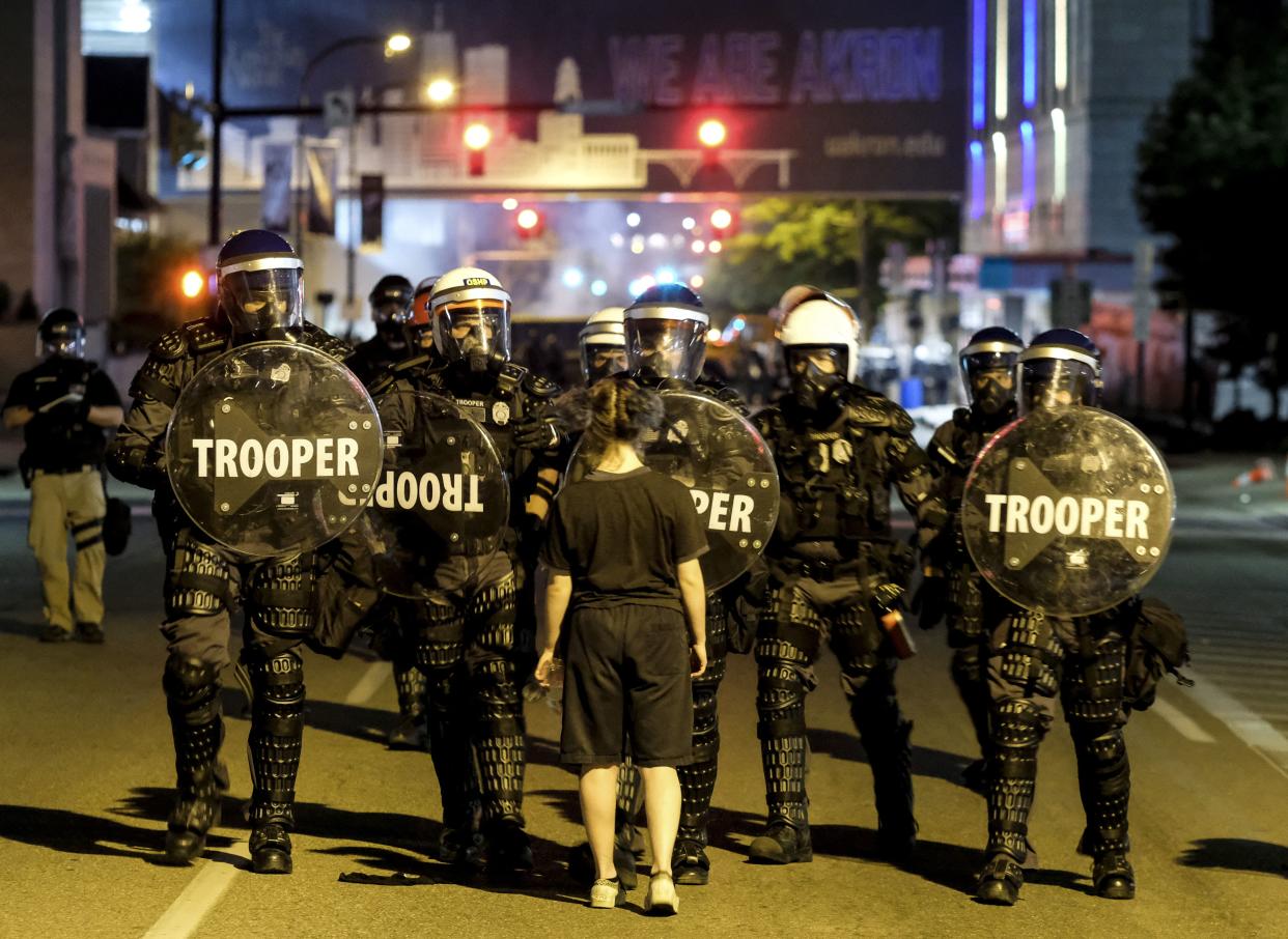 A woman stands in front of troopers in riot gear as police deploy tear gas and stun grenades.