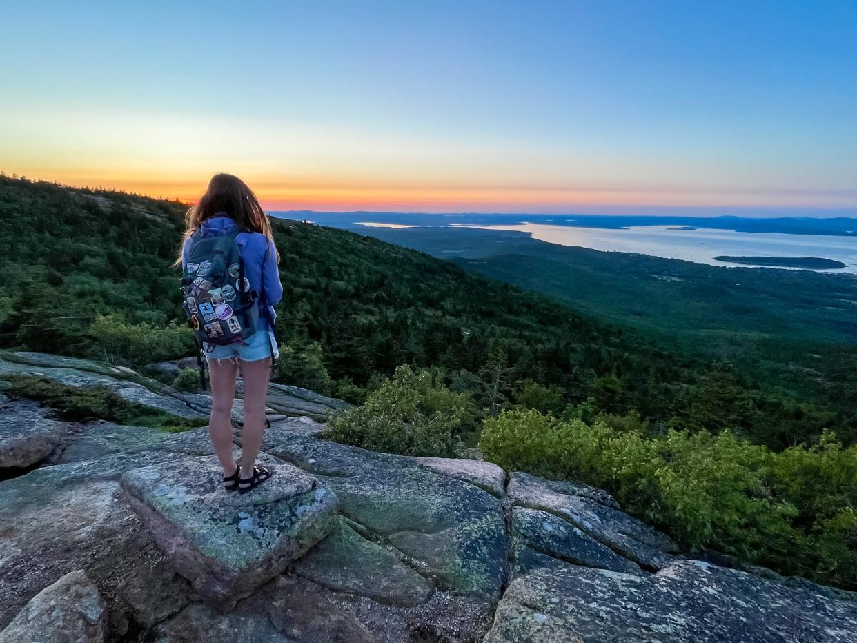 Emily standing on a rock formation facing a viewpoint of greenery and a body of water as the sun sets at Acadia National Park.