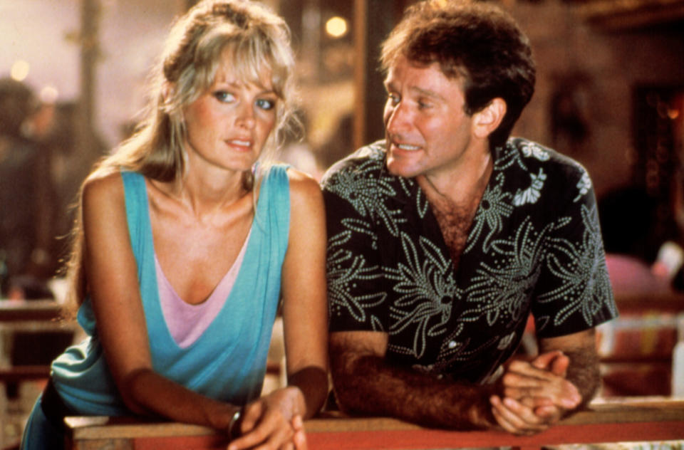 A scene from a movie featuring actors wearing casual clothes. The woman has a light-colored tank top and the man wears a patterned shirt, looking at her. Names unknown