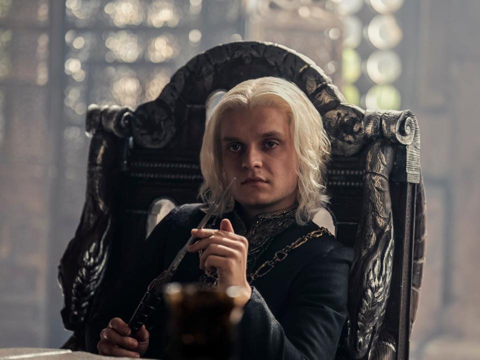 tom glynn-carney as aegon targaryen in house of the dragon. he's sitting in a high backed chair, playing with a steel dagger, and looking across a table