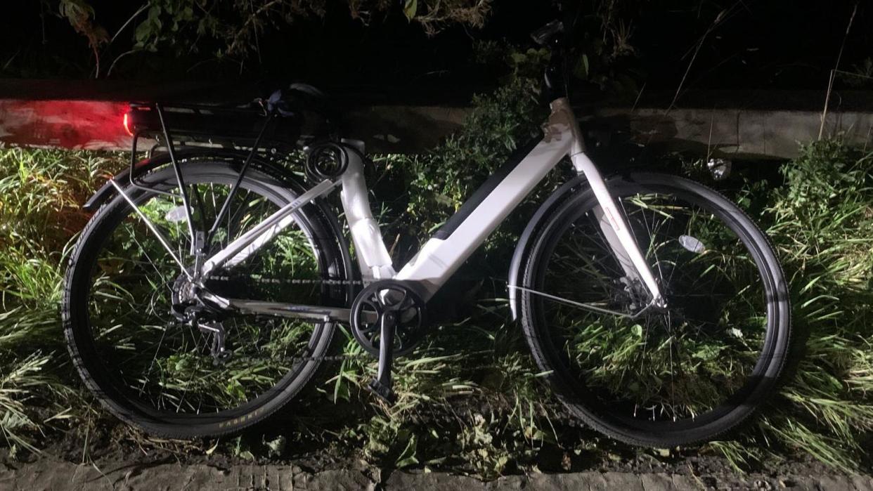 A modified e-bike lies on the ground, on grass on the hard shoulder of a motorway at night time. The wheels are black and the frame is silver in colour. The back break light is glowing red across the floor. 