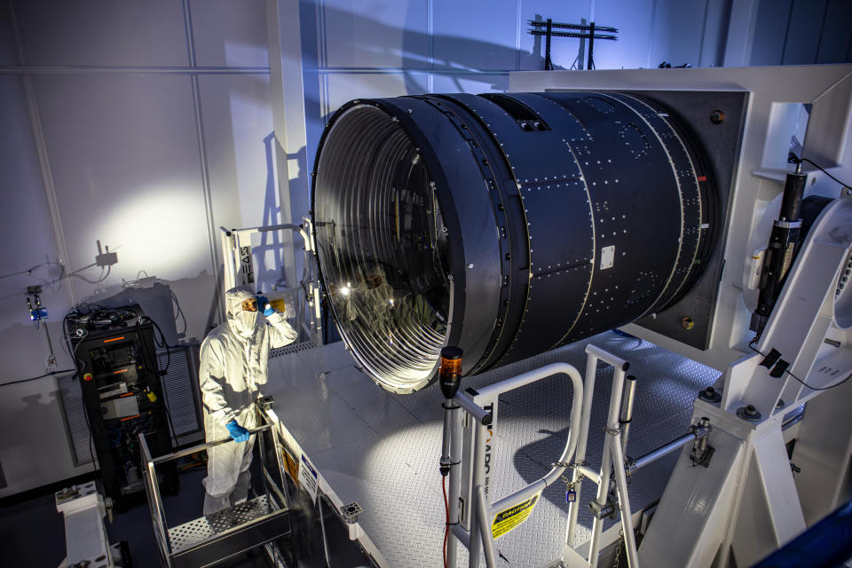 a small person in an all-white jumpsuit peers into a giant lens attached to metal supports in a white, dimly lit room.