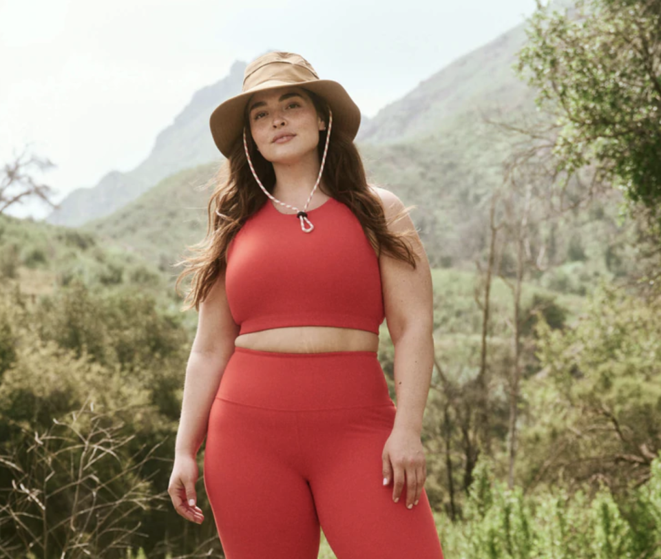 plus size model wearing red top, red leggings, and brown hat in mountains and forest, The Great + Eddie Bauer The Hiking Tank in Pimento (Photo via The Great)