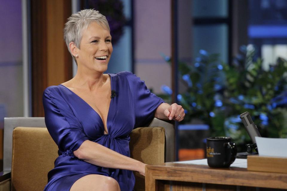 jamie lee curtis wearing a purple dress, smiling and sitting at a chair next to a desk on a television set