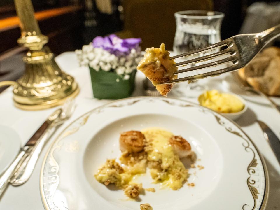 A fork holds a bite of scallop over the dish on a white plate