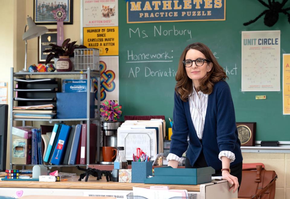 Tina Fey as Ms. Norbury in a classroom, leaning on a desk with books and papers, with a chalkboard behind her displaying math-related content