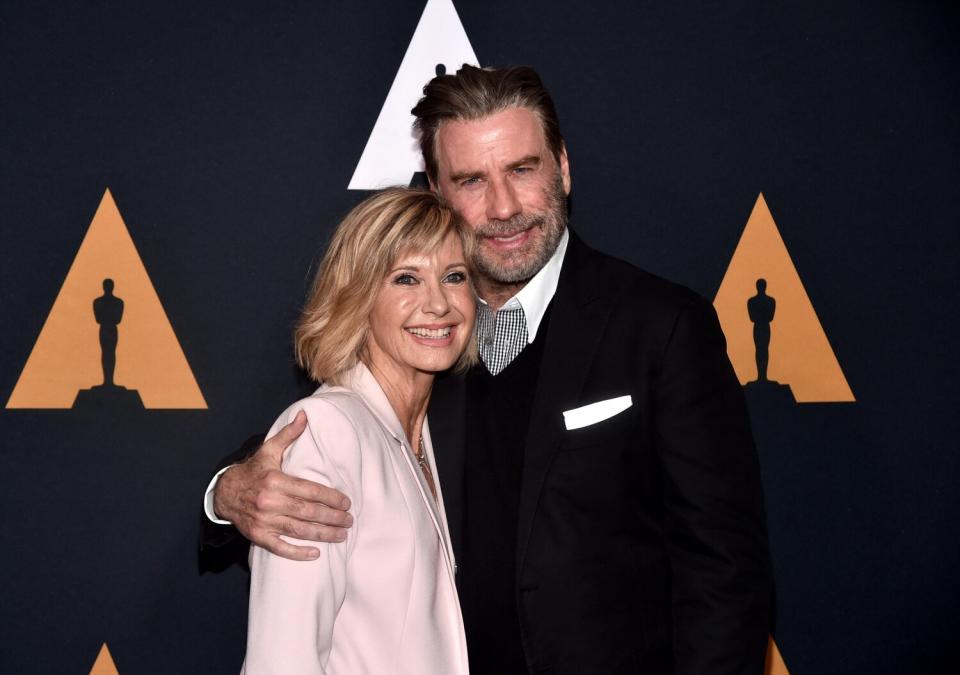 A man in a dark suit smiles and wraps his arm around the shoulders of a shorter woman wearing pink