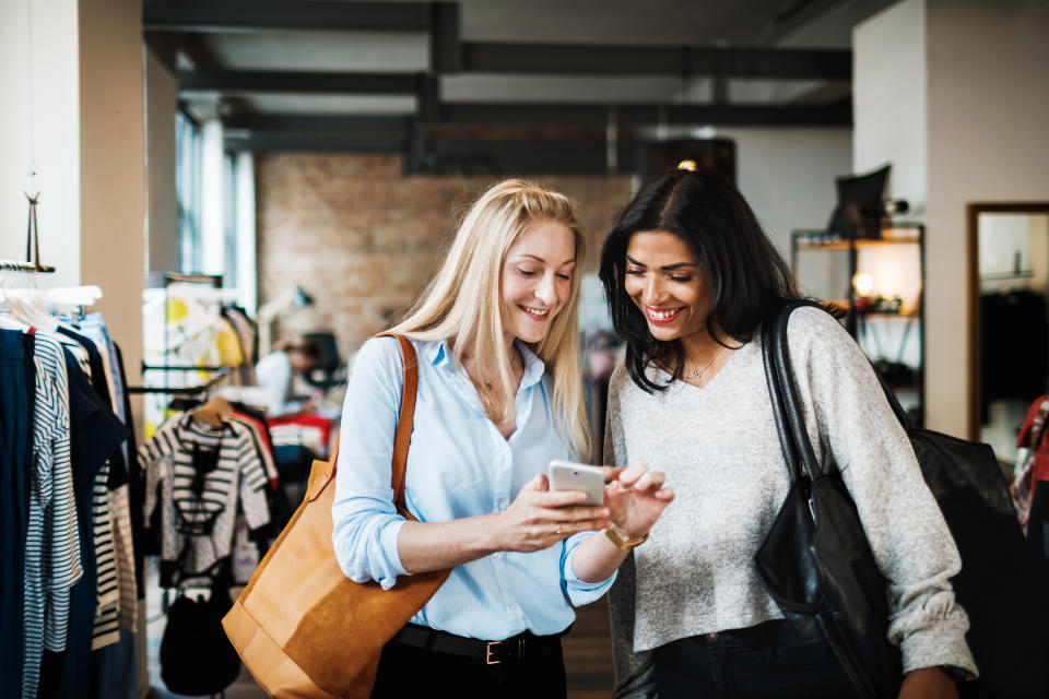 Two women smiling and looking at a smartphone while out shopping for the day together.