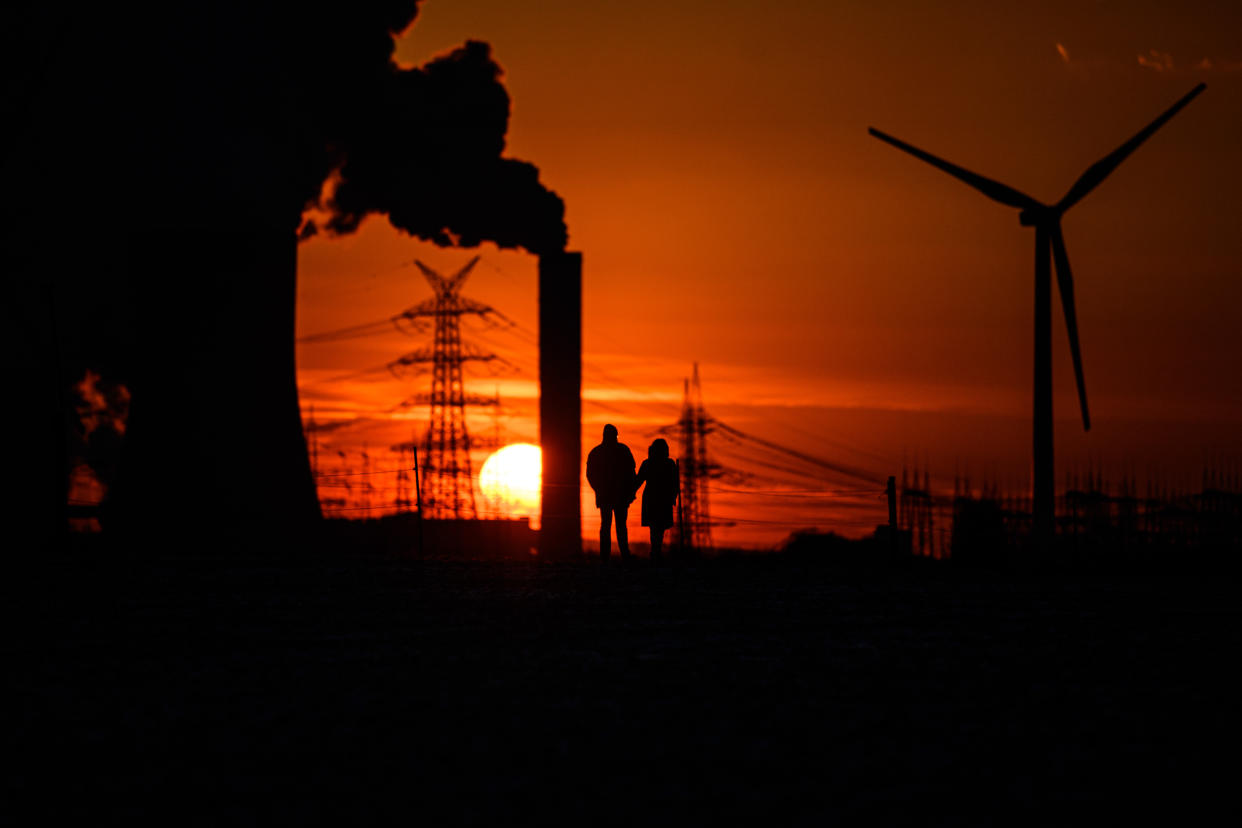 Two people stand before a sunset near steam rising from cooling towers of a power plant