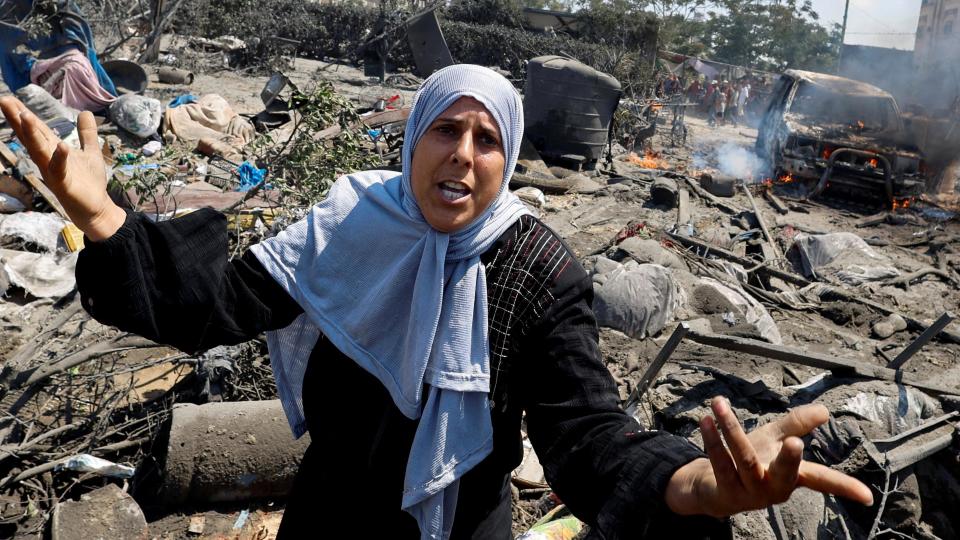 A woman raises her arms in fear. Behind her, the aftermath of an Israeli airstrike can be seen, including a burnt-out truck.