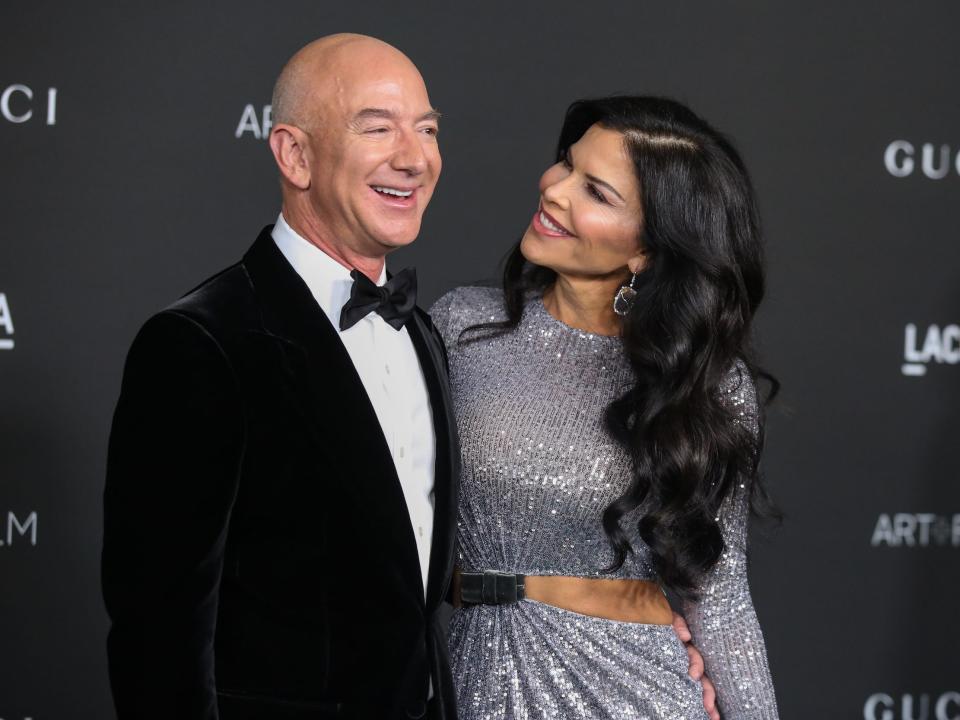 Jeff Bezos and Lauren Sanchez laugh while standing on red carpet against black backdrop