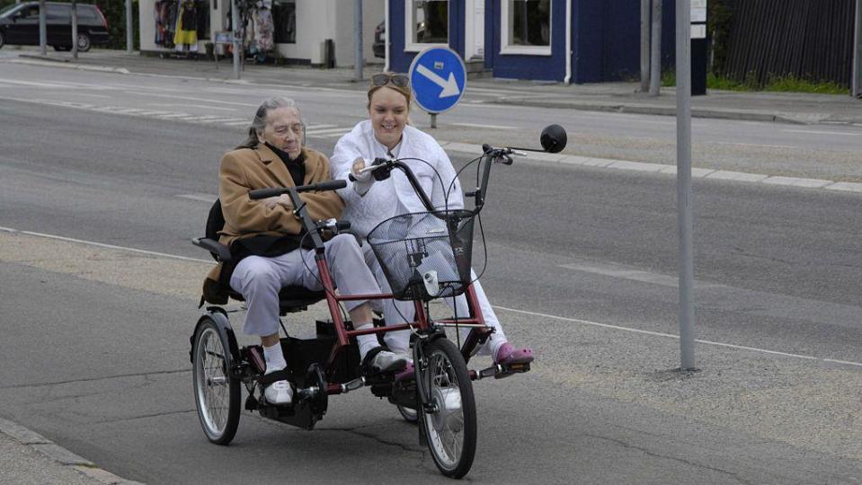 Una mujer anciana en una bicicleta junto a una asistenta de los servicios sociales.