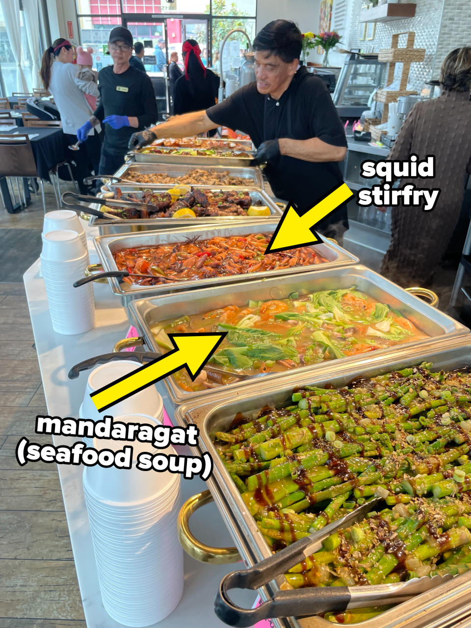 People serving and arranging a variety of dishes at a buffet with multiple food trays filled with assorted foods and sauces