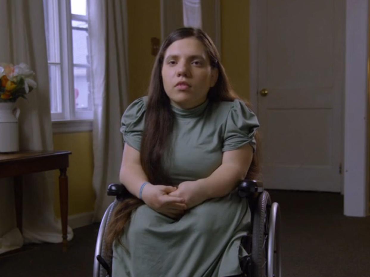 natalia grace barnett, wearing a green dress, her hair long and brown, and sitting in a purple wheelchair in a yellow painted room