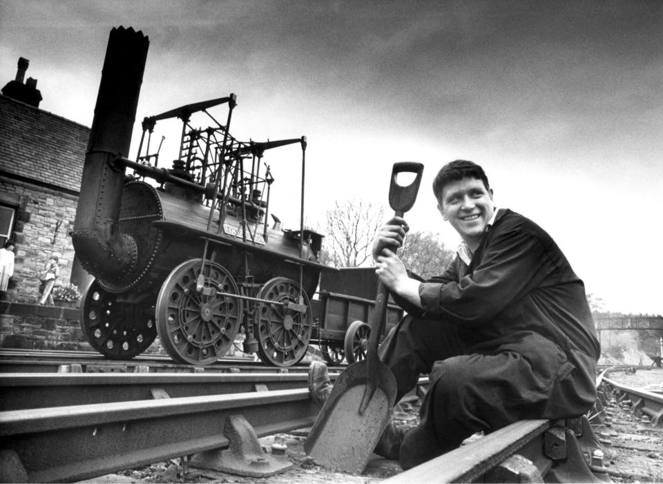 A man holding a shovel sitting in front of an old locomotive.