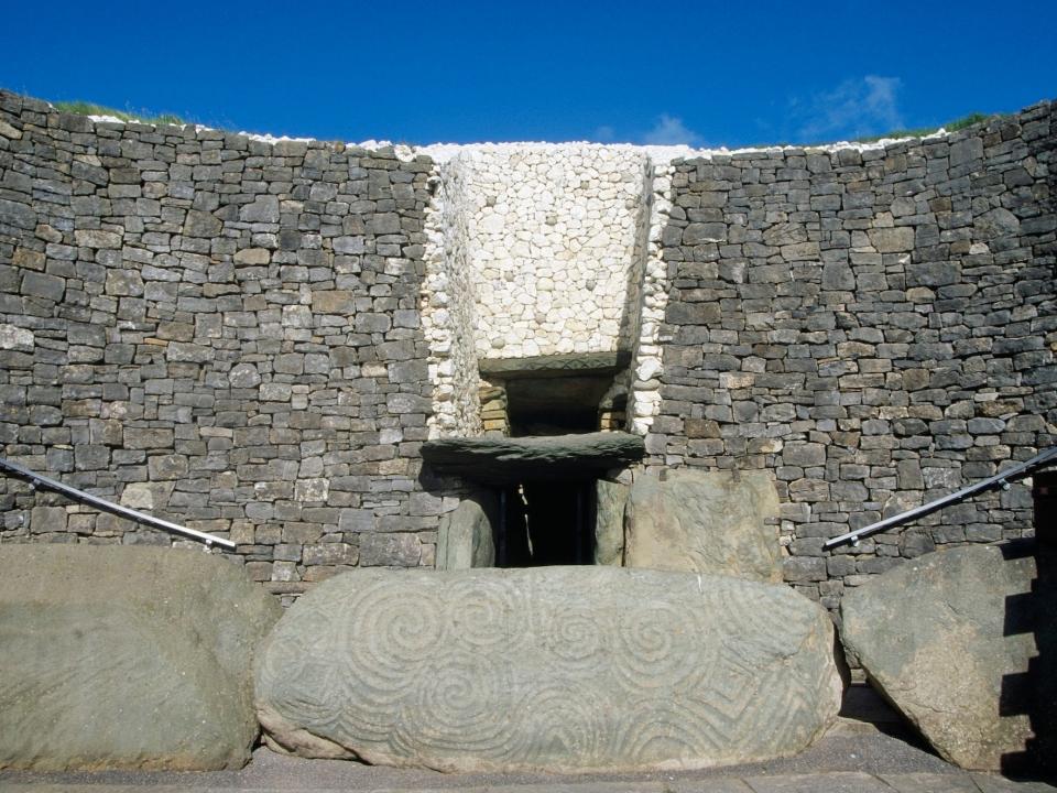 A picture shows the entrance of Newgrange. An opening peers between monolythic walls of grey stone stacked on top of each other. In fron of the opening is a large boulder carved with celtic shapes.