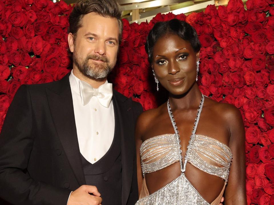 Joshua Jackson, in a black jacket with tails, and Jodie Turner-Smith, in a sparkly silver bodysuit, pose in front of a background of roses at the 2022 Met Gala.