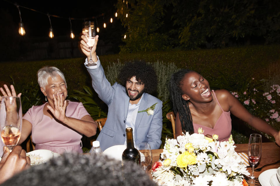 Three people at a celebration, one man and two women, are smiling and raising their glasses while seated at a table adorned with flowers and lit by overhead string lights