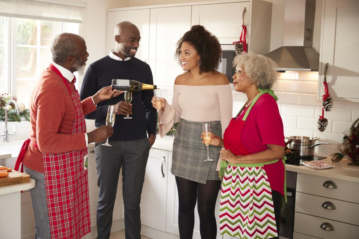 Young couple with two grandparents, grandfather is pouring champagne during a Christmas celebration at home in the kitchen, island is on the left with counter and windows in the background