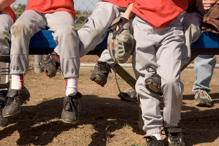 Children in baseball uniforms sitting on a bench, legs dangling, wearing cleats