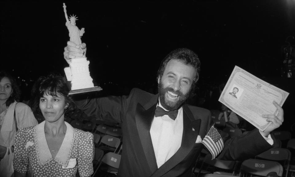 Yakov Smirnoff, smiling and in a dinner jacket, holds aloft a model of the Statue of Liberty and a certificate of his U.S. citizenship.
