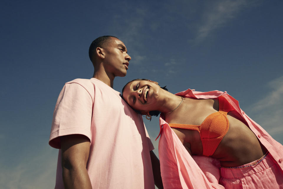 A man in a T-shirt and a woman in a bikini top and open shirt pose against a clear sky. The woman leans her head on the man's shoulder, both smiling