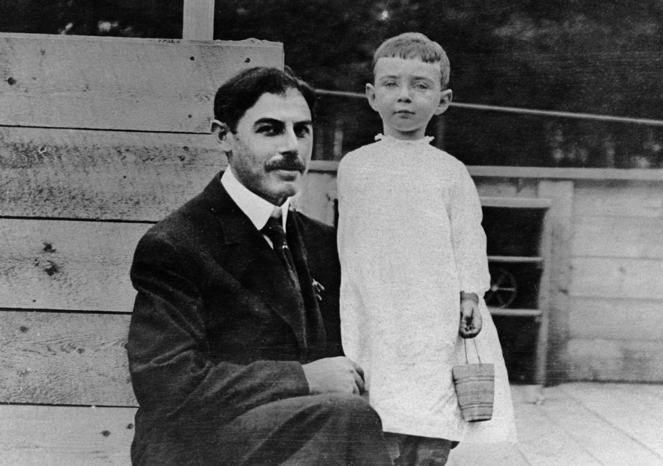 a man in a suit and tie kneels next to his son, who wears a white outfit and holds a bucket