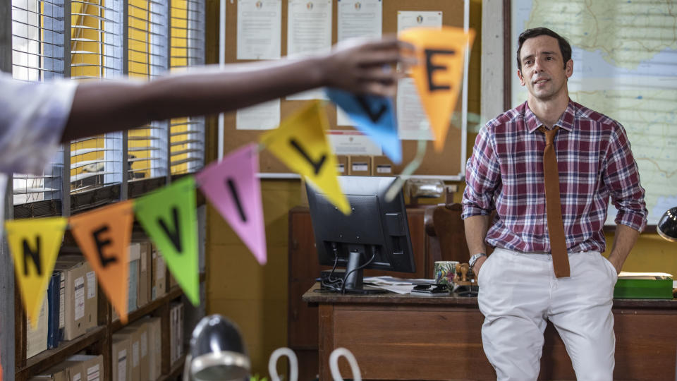 Neville (Ralf Little) is sitting on the edge of his desk in the police station and smiling. In the foreground, Naomi (Shantol Jackson) - who is just out of shot - is holding up some bunting with Neville's name on it.