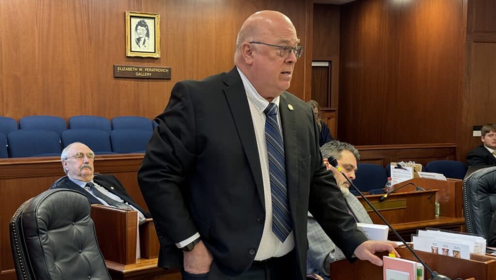 A bald, dark-suited man stands at a desk and speaks into a microphone