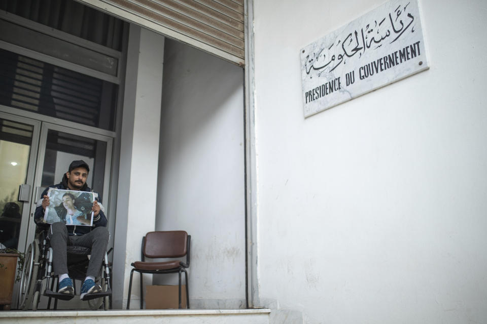 Rached El Arbi, 30, a protester who was paralyzed after being shot during Tunisia's democratic uprising 10 years ago, poses for a portrait while holding an image of himself, in Tunis, Tunisia, Tuesday, Jan. 12, 2021. El Arbi, now 30, has been paralyzed since being shot while protesting the autocratic regime of Tunisia's President Zine El Abidine Ben Ali, who was overthrown on Jan. 14, 2011. A photo of El Arbi hospitalized at the time went viral across the Arab world. (AP Photo/Mosa'ab Elshamy)