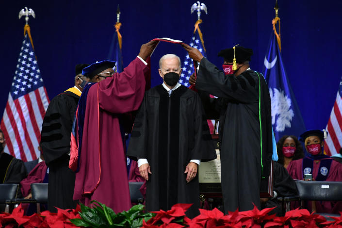 President Biden stands onstage during a commencement ceremony with others, all wearing robes and face masks.