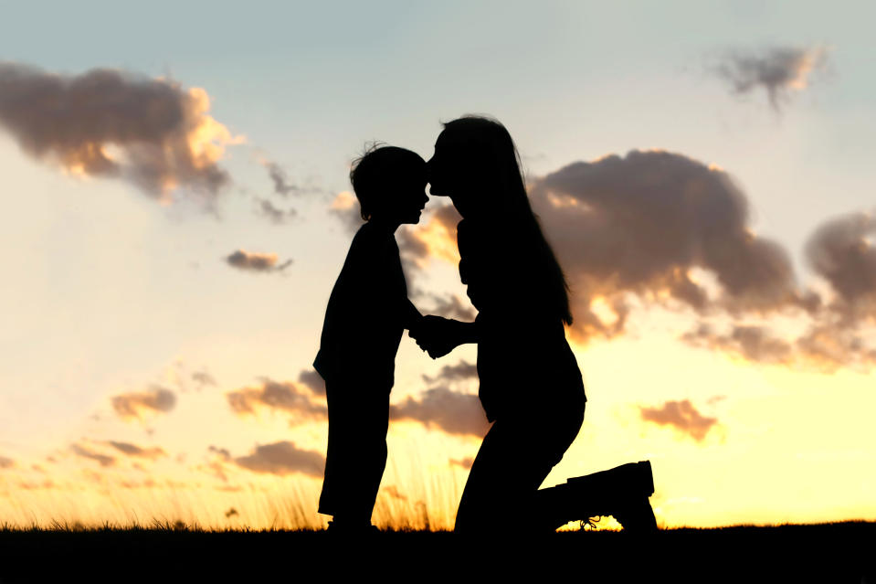 Silhouette of a young mother lovingly kissing her little child on the forehead, outside isolated in front of a sunset in the sky.