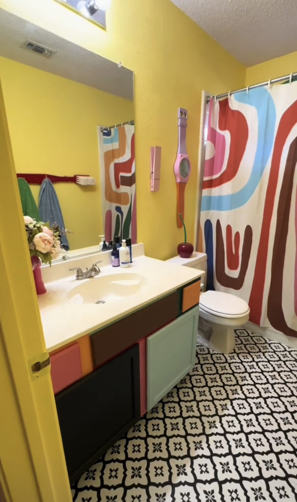 A colorful bathroom with an abstract-patterned shower curtain, a multicolored cabinet, and a geometric-tiled floor, featuring a vanity with various toiletries