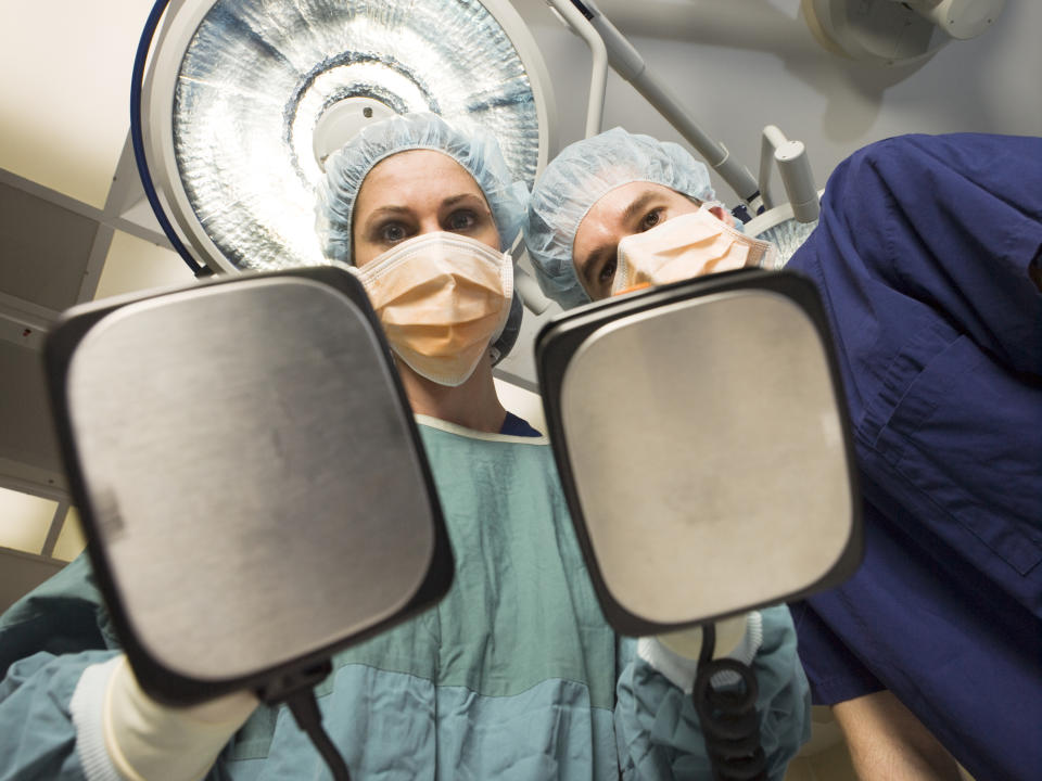 Two surgeons in scrubs and masks in an operating room, holding defibrillator paddles, looking down at the camera