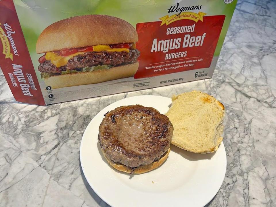 A green and red box of Wegmans premade burgers with a cooked burger on a bun with the top of the bun placed next to it on a white plate