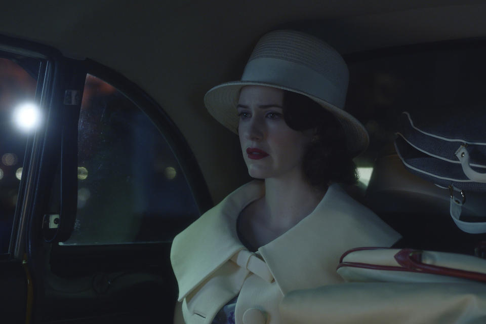 A woman in a white 1950s coat and hat sitting next to a stack of luggage in the back of a taxi; still from "The Marvelous Mrs. Maisel."