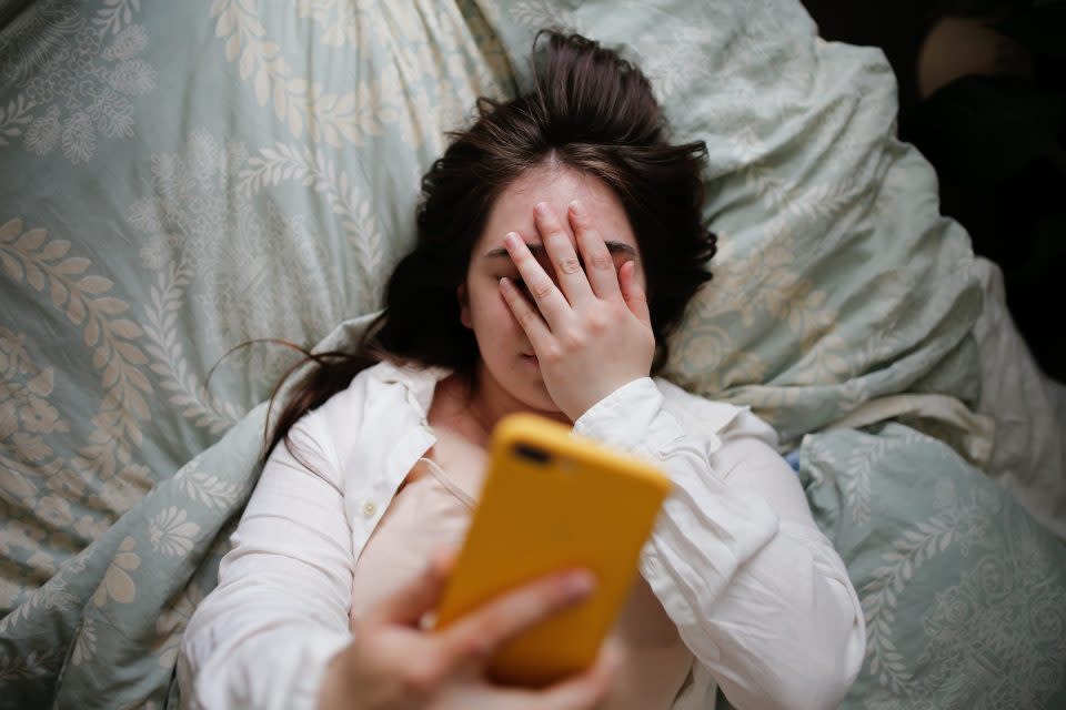 Sleepy young woman with flowing hair using phone on bed, close face palm. Top view