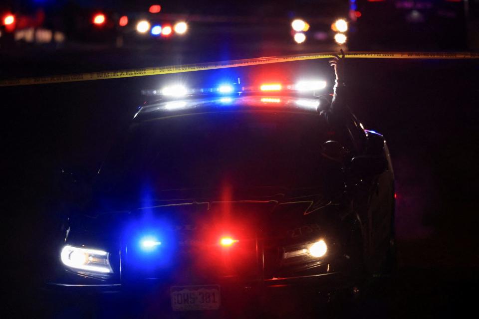 A Colorado Springs Police officer lifts a yellow cordon in front of a police vehicle at night as they attend the scene of a shooting at LGBTQ club Club Q early on November 20, 2022