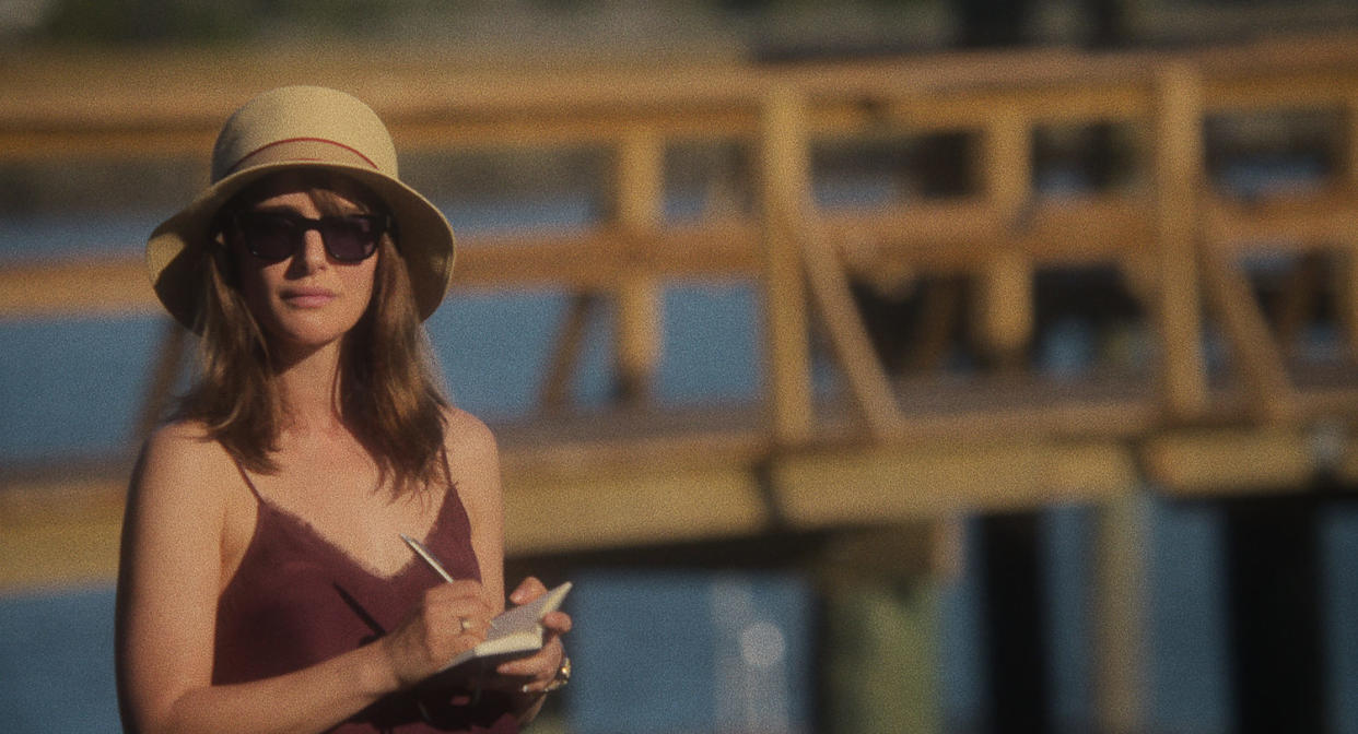 Elizabeth stands in front of a dock holding a notepad and pen, wearing a purple dress and wide brim hat (Courtesy of Netflix)