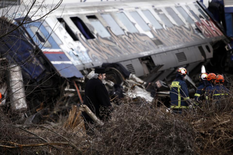 A Greek Orthodox priest stands near firefighters as they operate after a collision in Tempe, about 376 kilometres (235 miles) north of Athens, near Larissa city, Greece, Wednesday, March 1, 2023. Rescuers searched Wednesday through the burned-out wreckage of two trains that slammed into each other in northern Greece, killing and injured dozens of passengers. (AP Photo/Giannis Papanikos)