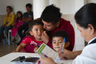 In this Jan. 28, 2020 photo provided by Cronkite News, Arizona State University, Agustin Reyes of Guererro, Mexico, wipes tears from 3-year-old Edgar Reyes at Casa del Migrante before receiving shots and medical care in San Luis Rio Colorado, Mexico. One Hundred Angels, a Phoenix organization that provides medical care and other services to migrants, helped coordinate the one-day vaccination clinic at Casa del Migrante, working with the Mexican Red Cross. (Delia Johnson/Cronkite News, Arizona State University via AP)