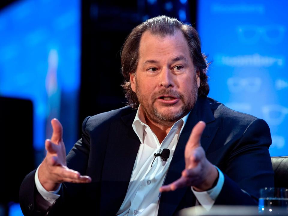 Salesforce founder Marc Benioff, wearing a sports coat jacket and in front of a blue background on a stage, extends his hands forward during a talk.