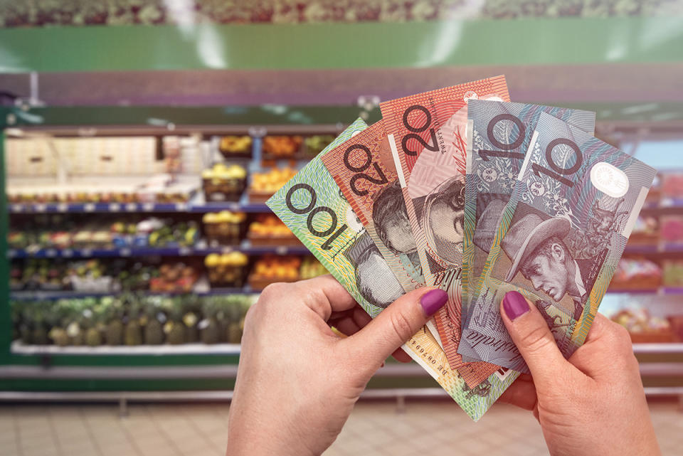 Hands holding Australian money in front of a Woolworths fresh produce section.