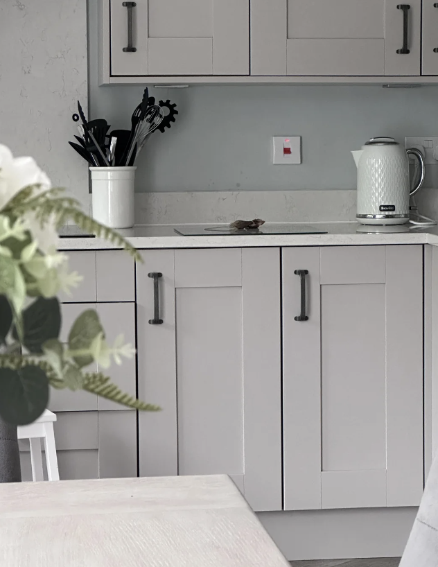 Modern kitchen with light cabinetry, utensils in a holder, an electric kettle on the countertop, and a light switch. There is a hint of flowers in the foreground