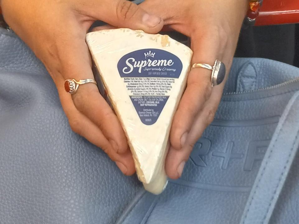 hand holding a wedge of brie cheese over a shopping cart at trader joe's